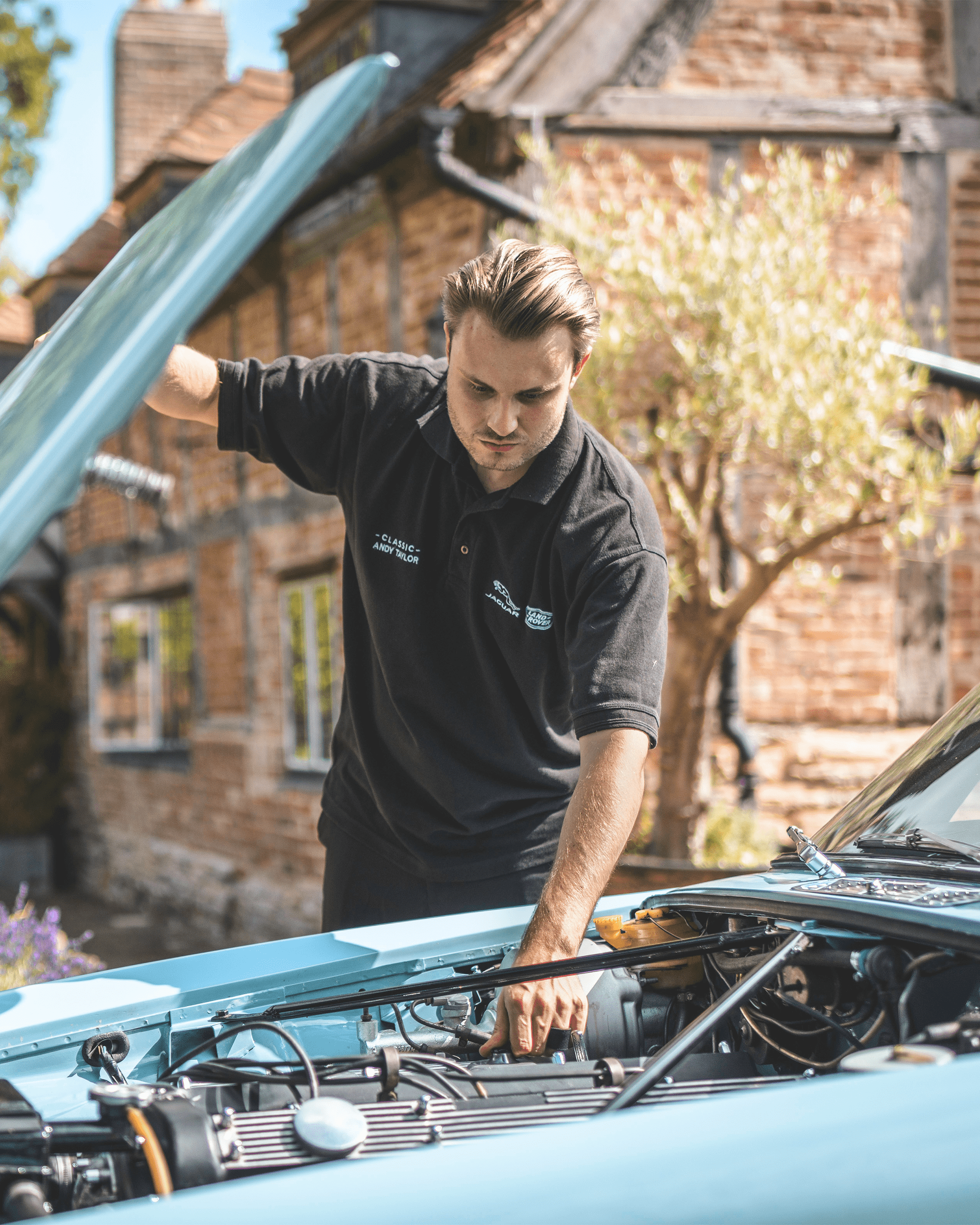 Mechanic working on engine of light blue car with open hood, outside brick building.