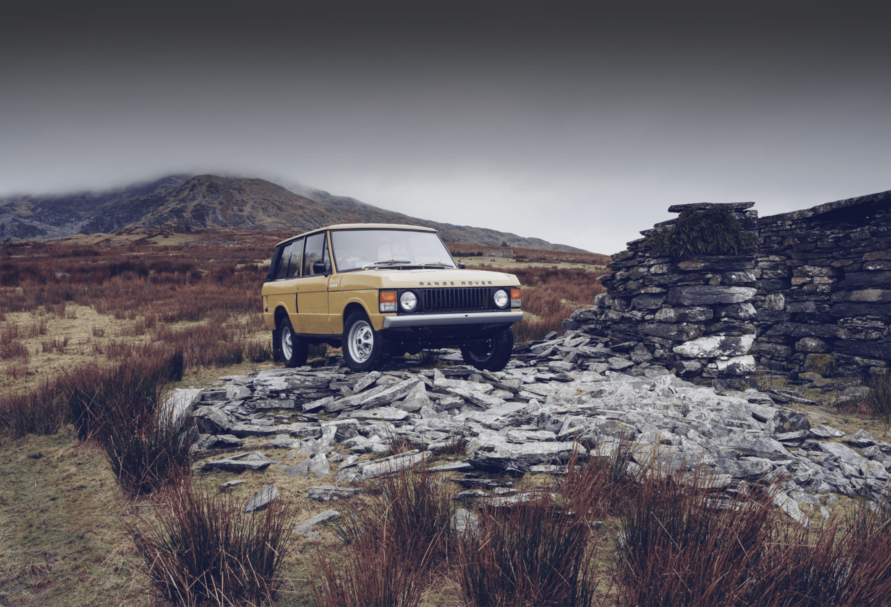 Yellow Range Rover on rocky terrain beside stone structure, with hills in background.