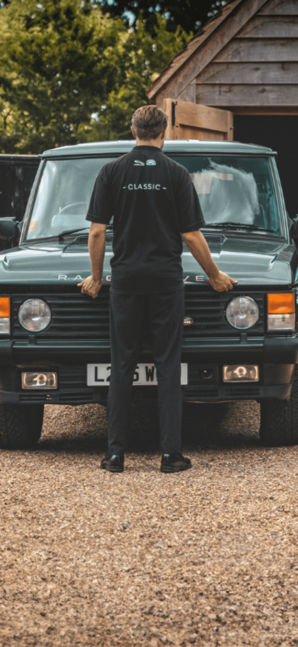 Mechanic in “CLASSIC” uniform standing by green Range Rover near open garage with blue classic Jaguar inside.