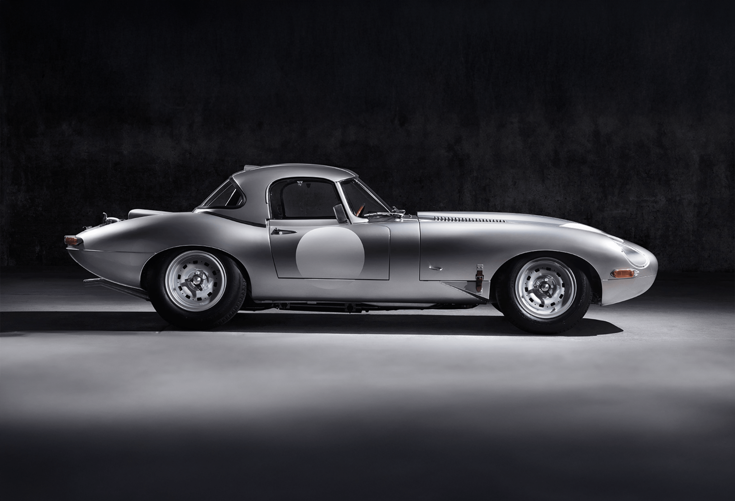Classic silver E-Type with racing circle on door, side view.